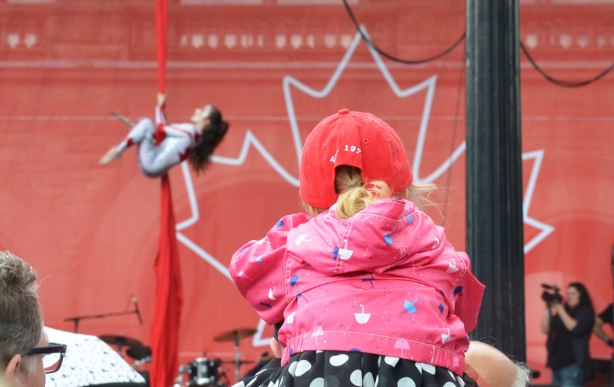 The back of a young girl wearing a red baseball cap and sitting on someone's shoulder as she watches an acrobat show on a stage