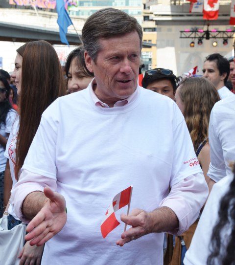 Mayor John Tory has a small Canadian flag in his hand as he talks to people at Dundas Square on Canada day.   He's in a white T-shirt. 