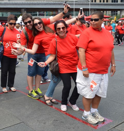Five people pose for a photo.  They are wearing red T shirts and they are standing withing the stem of the maple leaf outline that is taped to the ground.  They are the first ones into the living flag that is trying to be made at Dundas Square as part of a Canada Day celebration 