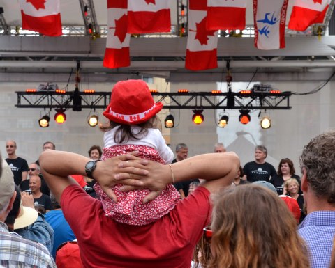 A man holds a young girl on his shoulders while the watch a performance of New Choir, a choir that sings old rock songs, as part of a Canada Day celebration at Dundas Square.  The girl is wearing a red hat with white maple leafs on it.  There are Canadian flags hanging from the ceiling of the stage. 