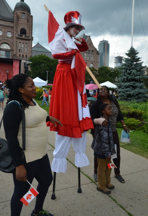 A person in red and white Canada Day costume is on stilts with three others standing beside, in front of the parliament buildings at Queens Park 