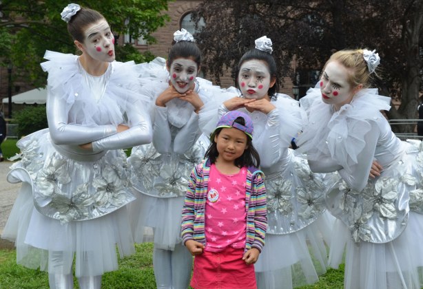A young girl looks a bit skeptical as she poses with four women dressed in frilly white dresses who are hamming it up for the camera.  Exaggerated facial expressions