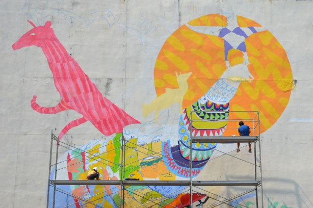 Two people sitting on scaffolding while they paint a mural