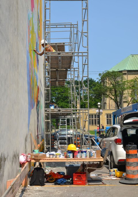A man wearing a hard hat who is sitting on scaffolding and painting a mural on the side of a building. 