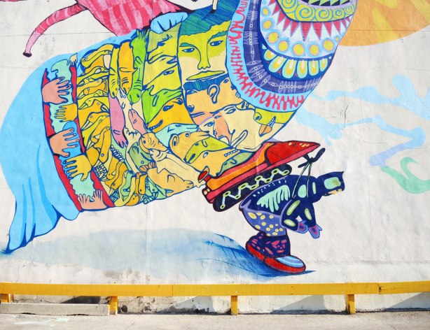 Close up picture of the mural's bottom center part showing a man in winter clothing with a snowmobile strapped to his back.  He is hunched over as he walks.  On the snowmobile of a very large bird shaped crature with faces covering its body. 