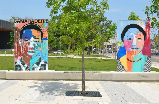 on a concrete plaza at the entrance to a park, large glass laminate artwork that is a colourful portrait of a person - two people, on the left is a young man with the word camaraderie and on the right is a young woman with the word appreciate