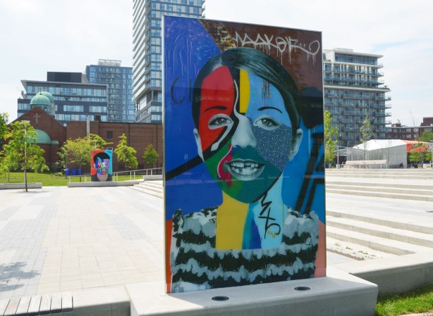 on a concrete plaza at the entrance to a park, large glass laminate artwork that is a colourful portrait of a person - a boy