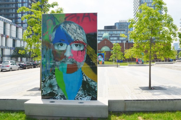 on a concrete plaza at the entrance to a park, large glass laminate artwork that is a colourful portrait of a person - 