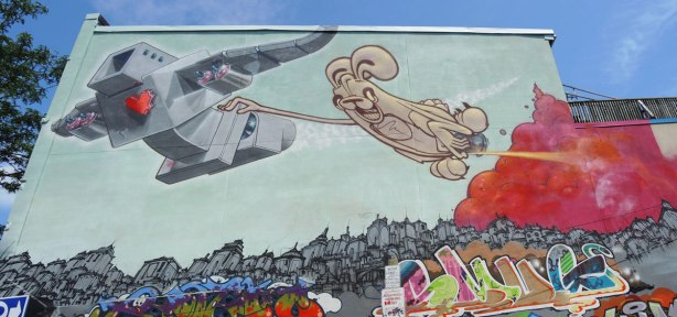 a mural on the upper storey of a two storey building.  Lovebot is flying above a city scene with a light brown poser bunny holding on to him, and flying behind him