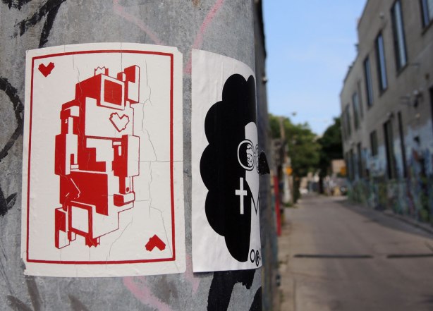 graffiti and street art in Graffiti Alley in Toronto - a lovebot of hearts playing card sticker on a metal pole beside a sticker of a woman's head with curly black hair and big earrings in the shape of a cross
