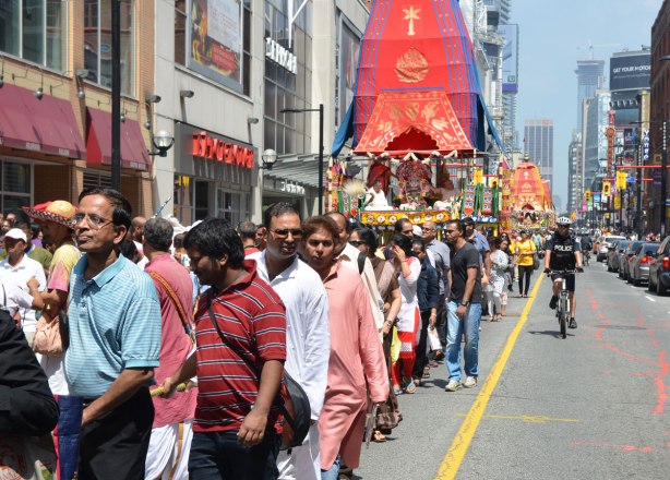 parade going down Yonge St. using just the southbound lanes. A policeman on bike keeps the traffic out of the way. A float is being pulled by people walking in the parade. Part of the Festival of India in Toronto.