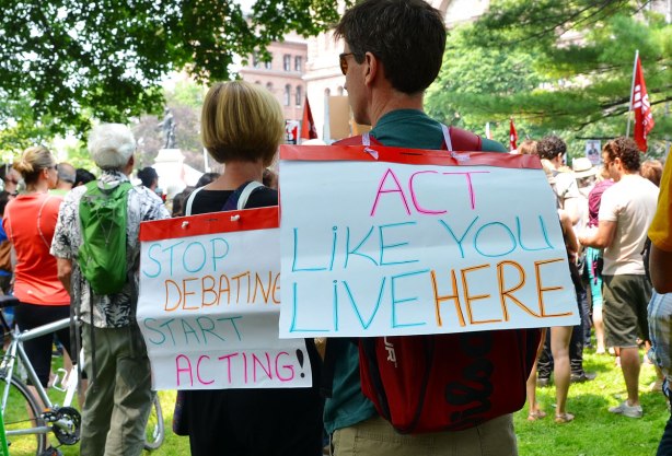 Two people with protest signs on their backs.  One says 'Act like you live here' and the second says 'Stop Debating, Start acting'