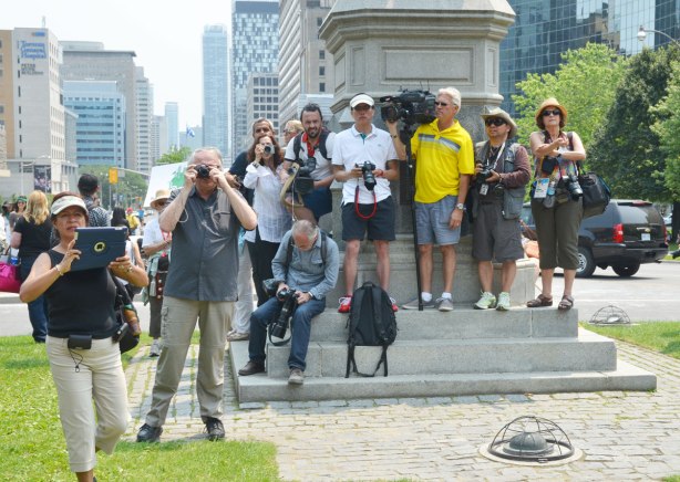 Many photographers are standing on the base of a statue while they wait for a protest march to begin in Toronto at Queens Park 