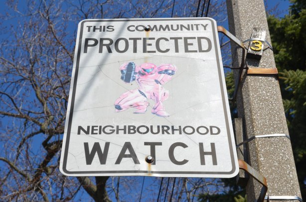 An altered Neighbourhood Watch sign. A picture of a superhero dressed head to toe in pink (or is it faded rad?) and holding a large weapon has been added to the sign