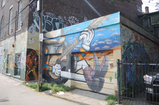 graffiti and street art in Graffiti Alley in Toronto -  Two buildings in the picture.  In the foreground is a one storey garage with a large totund stylized guitar player sitting on the ground with one knee up.  Behind him you can see another painting of a guitar player but this one is a bit more realistic. 