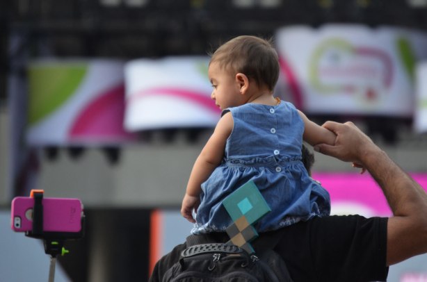 A toddler sits on her father's shoulders.  He is holding onto one of her arms.  She is looking at a pink smartphone being held up by a selfie stick almost to her level. 