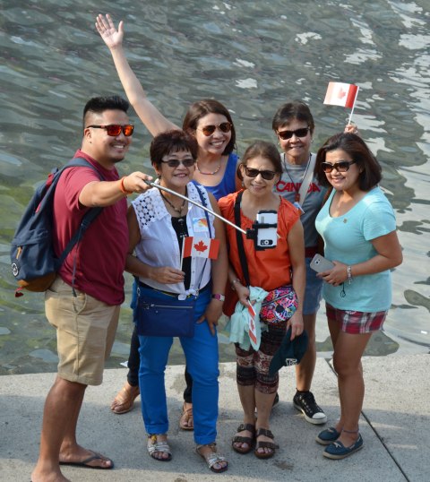 Six people, a man and five women, are talking a selfie with a phone on a selfie stick.  Two of the women have small Canadian flags.  Another woman has her arm up in the air. 