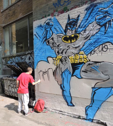 A young man is spray painting part of a batman mural.  The figure of batman is completed and is about 4 meters tall.  A city scene is being added in the bottom corner