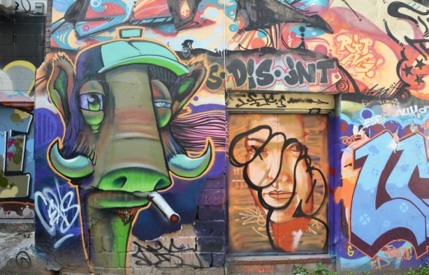 A stylized greenish brown bull with horns is smoking a cigarette, only the head is shown.  It is on a wall and beside it is a door that had a woman's face painted on it.  The face has been tagged over.   Dis jnt is written across the top of the door.  disjoint perhaps.
