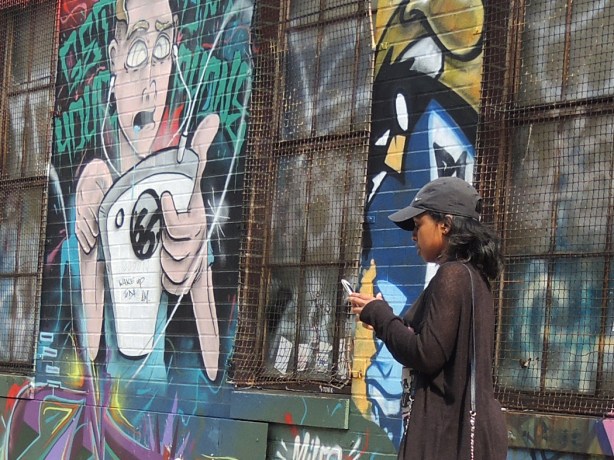 Woman taking picture on a smartphone, standing beside a street art piece of a man with earphones plugged into 