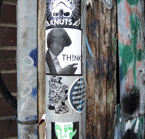 graffiti in an alley - 4 stickers on a metal pole. Knuts on the top, a woman in profile with the word think next, black and white drawing third and on the bottom is a green on white line drawing of a man's face