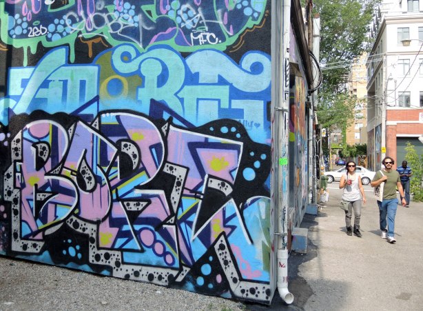tags and letter graffiti in blues and purples on an alley wall, with a few people walking down the alley as well 