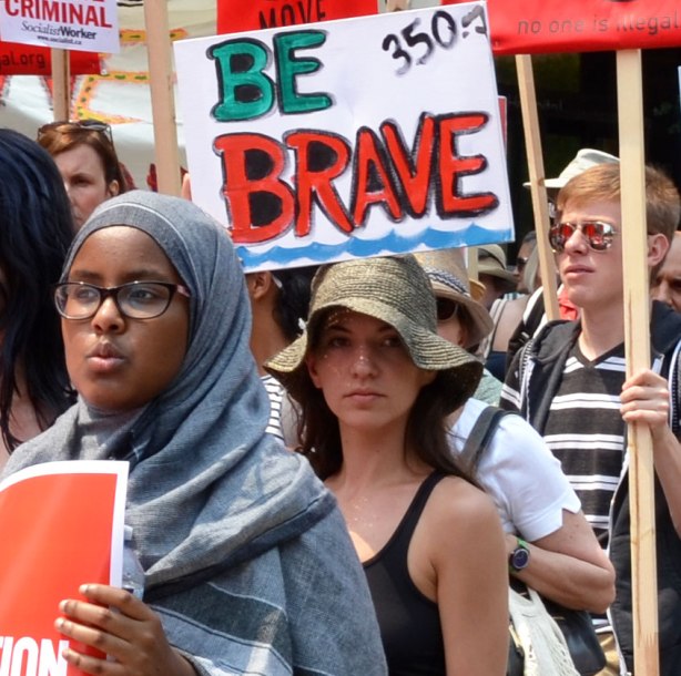 jobs, justice and climate action protest march  - a young woman in a head scarf holds a red and white sign, the woman behind her is wearing a floppy hat and there is a sign that says Be Brave in large capital letters above her head. 