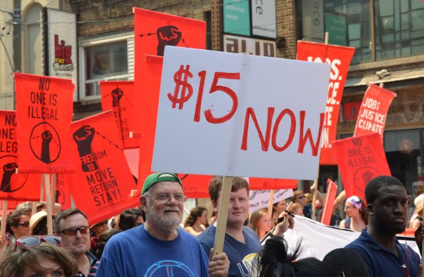 jobs, justice and climate action protest march -  a man is holding a sign that says $15 Now.  It is white with red letters.  It stands out because most of the signs around him are all bright red. 