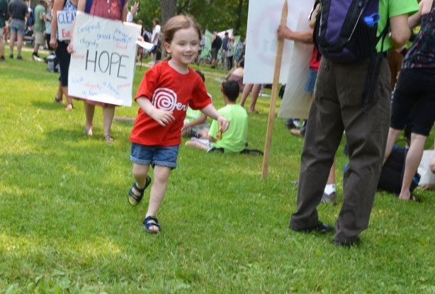 A young girl in a bright red Tshirt runs in front of some protesters, one of whom is holding a sign that says Hope. 