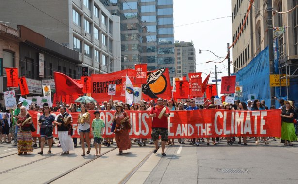 jobs, justice and climate action protest march - the lead banner of the march.  It says Jobs, Justice and the Climate. It is red with white letters and is almost the full width of the street.  A few drummers and dancers are in front of the banner.  Many people with signs and flags are behind the banner. 