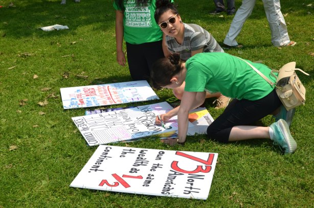 Three young women are making signs to carry in the protest march.  Two are in green T-shirts.  One of the signs says '73 North Americans own the same as 1/2 the continent.'  Jobs, Justice and Climate protest rally and march. 