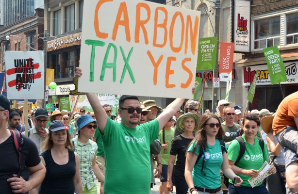 A man wearing a green party T-shirt is carry a large sign that says 'carbon tax yes'.  There are other green party supporters with him as they walk in a protest march in Toronto