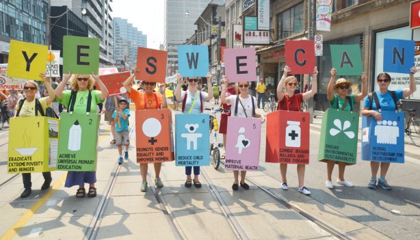 A number of women in a protest march, each is holding a different coloured sign and each has a different letter - together they spell 'yes we can'.  Each women is also wearing a coloured box that matches the colour of her sign and each box represents a differ aspect of social policy such as gender equality, primary education, poverty eradication, healthcare, 