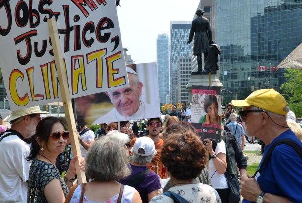 jobs, justice and climate action protest march -  a group of people at Queens Park, the back of the statue of Sir John A. Macdonald can be seen.  One woman is holding a sign that says Jobs Justice Climate.  Another person holds up a picture of Pope Francis.  They are all getting ready to march down University Ave. 