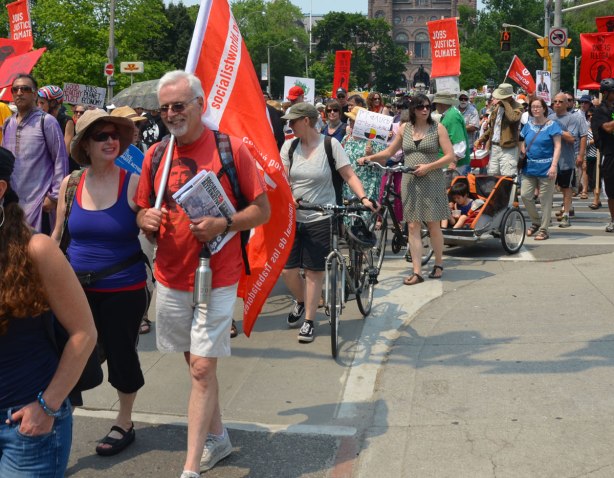 jobs, justice and climate action protest march - some of the people marching, a white haired man is holding an orange flag that says Socialistworld.net  Some people are walking their bikes. 