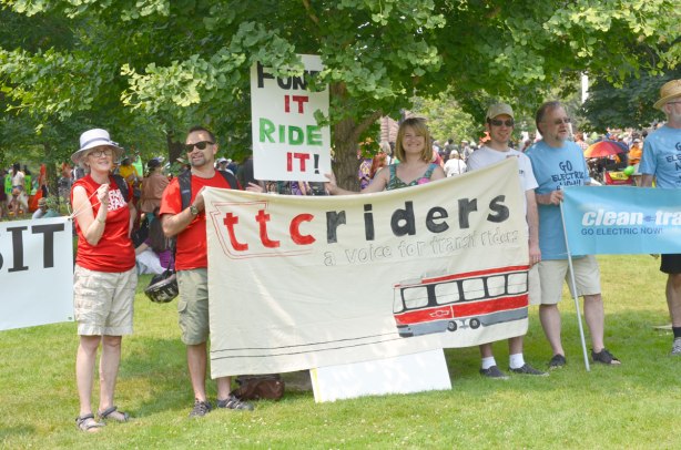At the jobs, justice and climate action protest rally, four people hold a banner that says 'TTC riders, a voice for ttc riders' and it has a picture of a street car on it as well. 