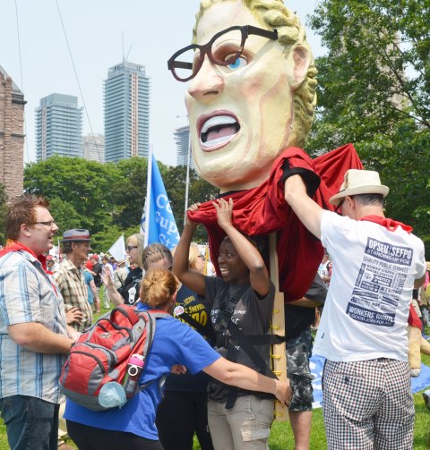 A couple of people are helping a woman prepare to wear a large effigy.  A very large head of a woman wearing dark rimmed glasses.  