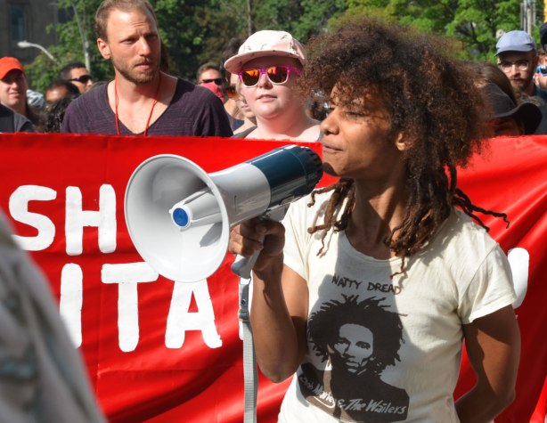 a young woman with afro black hair and wearing a Bob Marley T-shirts is speaking through a megaphone at a protest march.   She is standing in front of a red and white banner that is being held up by a few other people 