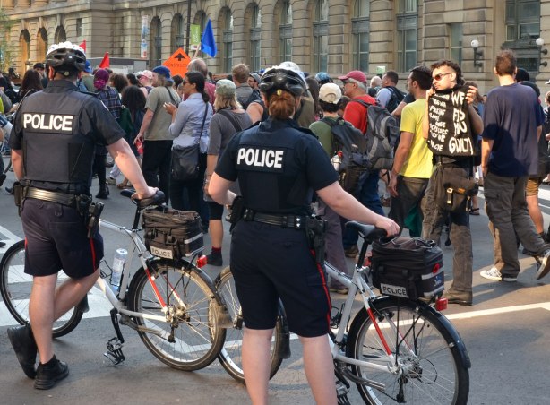 riseupTO demonstration and march - policemen and women stand besides their bikes as they do crowd control and direct the protest around a corner. A man is holding a black sign with white lettering that says "We find you guilty"