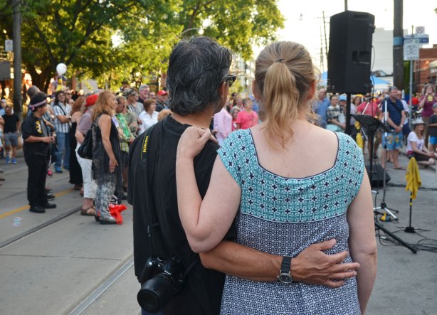 A man has his arm around a woman's back and her hand is on his shoulder as they watch a musical performance outside along with a number of other people