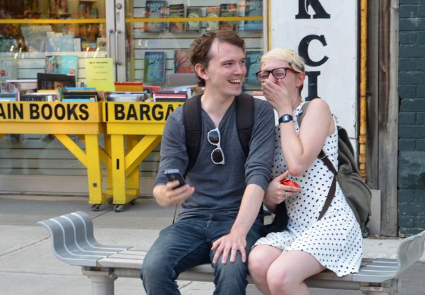 A couple sitting a bench laughing.  They were taking a selfie when they noticed 4 photographers taking their picture.  She is embarrassed and is covering her face, he is laughing. 
