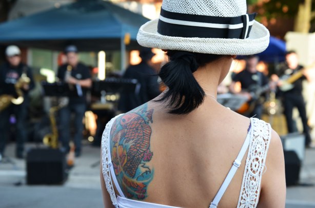A woman's back is to the camera as she watches live music outside.  Her black hair is tied in a short ponytail, she is wearing a white hat and a white sundress.  She has a large tattoo on one shoulder blade. 