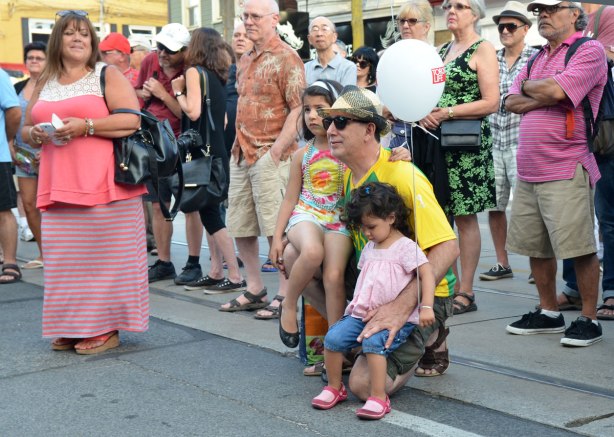 A man is squatting on the sidewalk with two girls on his knee. Both girls have balloons. They are part of a crowd watching a band perform in a street music festival