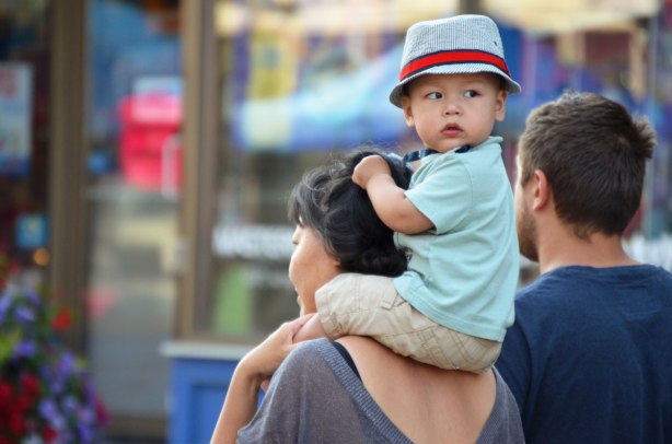A young Asian boy is on his mother's shoulders. He is looking back. He is wearing a greyhat with a red band, as well as a tie.
