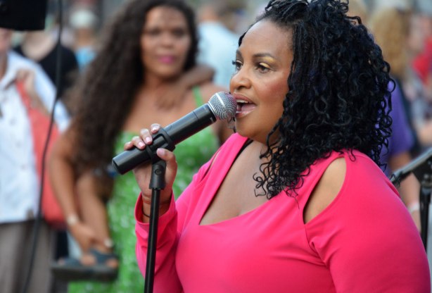 A black woman with shoulder length curly hair and a bright pink top is singing into a microphone. A woman in green is watching her