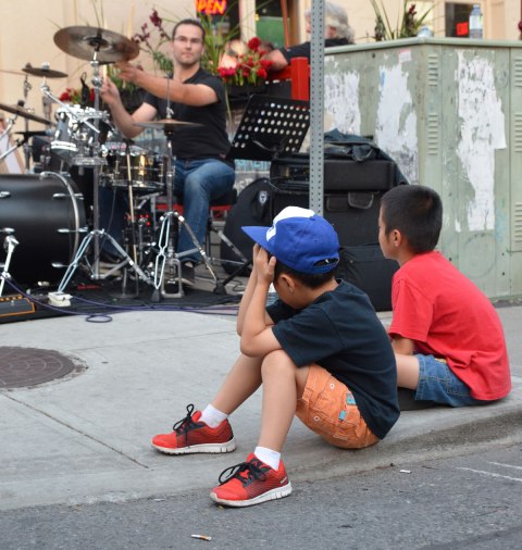 two young boys sitting on the sidewalk watching a drummer play in a band in a street music festival performance
