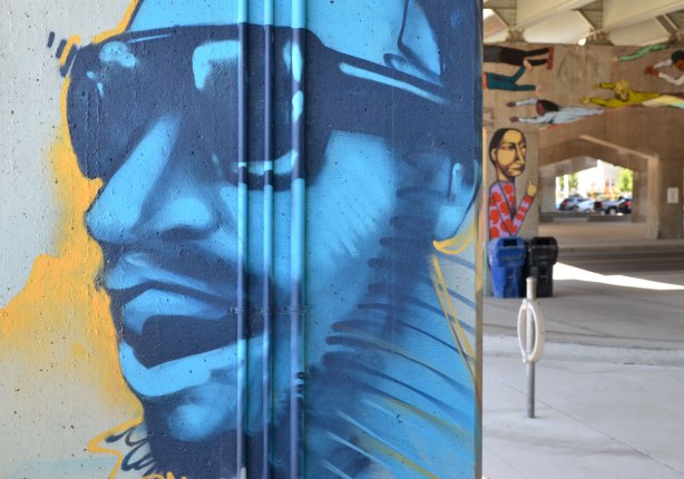 picture of art (mural) on a concrete support holding up a road above a skateboard and basketball park - large blue face of a man wearing sunglasses, in profile, in the foreground.  A pillar with a tall skinny person painted on it in the background. 