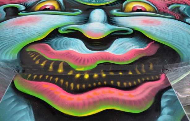 photo of art (mural) on a concrete support holding up a road above a skateboard and basketball park - close up of a multocoloured clown face with a scary expression