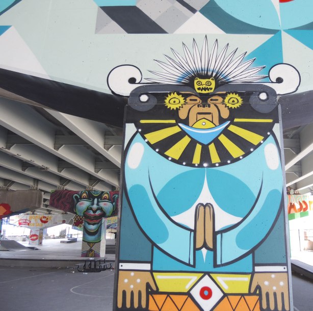 South American themed figure with head dress painted on a pillar, with other painted pillars in the background. 