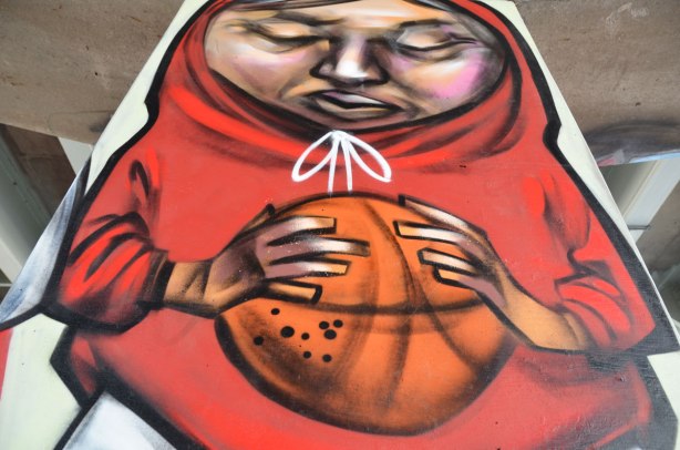 picture of art (mural) on a concrete support holding up a road above a skateboard and basketball park - an elicser painting of a person in a red hoodie holding a basketball.  Eyes closed, thining, hood pulled up over head 